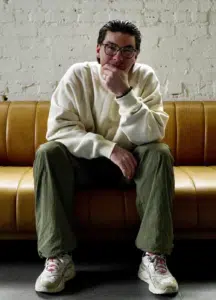A person wearing glasses, a white sweater, green pants, and white sneakers sits on a tan couch against a white brick wall, resting their chin on their hand.